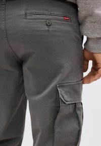 Γκρι cargo παντελόνι Levi’s με κουμπωτή πίσω τσέπη και τσέπη με καπάκι στο δεξί πόδι, φορεμένο από ένα άτομο που φοράει γκρι φούτερ.