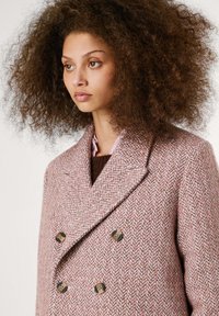 Manteau en laine rose en chevrons avec un design croisé, col en V, et boutons marron. Fini texturé avec des détails de couture visibles.