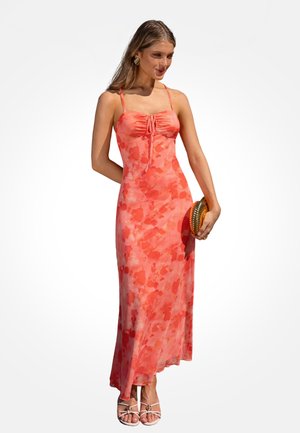 Femme portant une longue robe fleurie rose corail à fines bretelles, tenant une pochette ronde texturée et portant des sandales blanches à brides.