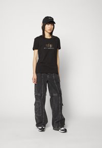 T-shirt noir avec le logo doré "Alpha Industries", associé à un pantalon cargo ample gris foncé avec des poches et des détails de couture.