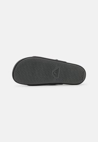 Semelle de chaussure en caoutchouc noir avec un motif de semelle ondulée et un logo, conçue pour l'adhérence et la durabilité avec une forme plate et large.