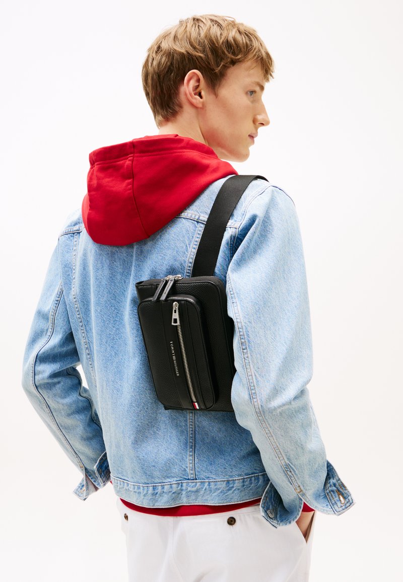 Černá křížová taška na zip s texturovaným materiálem, nošená přes světle modrou džínovou bundu s červenou mikinou pod ní.