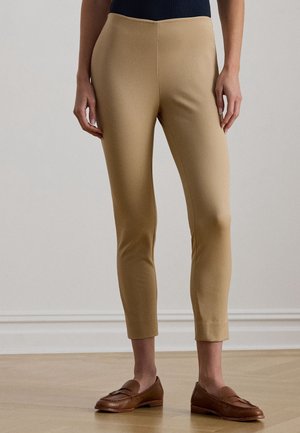 Slim fit beige cropped broek gedragen met bruine leren loafers en een donker topje, getoond van taille tot voeten op een houten vloer achtergrond.