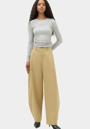 Donna con capelli lunghi e ricci che indossa una maglia a maniche lunghe grigio chiaro e pantaloni beige a gamba larga con stivali neri con tacco, in piedi davanti a uno sfondo semplice.