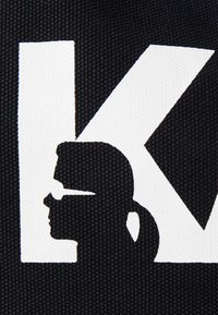 Tissu noir présentant le profil en silhouette blanche d'une personne portant des lunettes, à côté d'une grande lettre "K" blanche. Design de surface texturé.