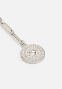 Porte-clés en argent avec un pendentif rond présentant un cœur en cristal au centre et les mots gravés "GUESS WITH LOVE FROM" autour.