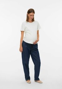 Mujer embarazada con una camiseta blanca y pantalones oscuros holgados, sosteniendo suavemente su barriga y mirando hacia abajo, de pie sobre un fondo blanco.