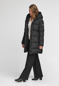 Czarna kurtka puchowa z kapturem, z suwakiem i guzikami, poziomymi szwami oraz naszywką z logo na rękawie. Połączona z czarnymi spodniami.