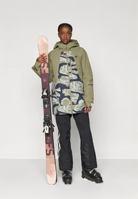 Slidinėjimo striukė alyvuogių žalios spalvos su kamufliažo raštais, derinama su juodomis slidinėjimo kelnėmis. Modelis laiko rožinius ir juodus slidžių su įtvirtinimais.