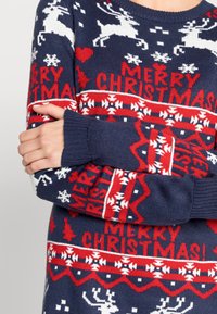 Marinblå stickad tröja med vita renar och röda accentfärger, med texten "GOD JUL" och snöflinge mönster genomgående.