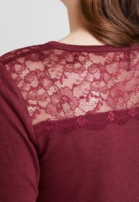 Bordeaux topp med en bakpanel av blommig spets med scalloped kanter, mjuk textur och rundad halsringning, som kombinerar avslappnade och eleganta designelement.