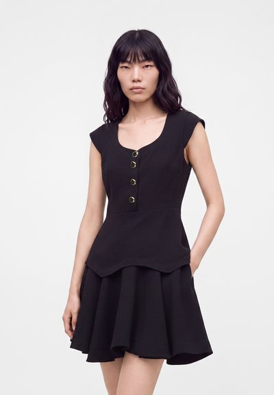 Femme aux cheveux noirs ondulés portant une robe noire sans manches avec des boutons dorés et une jupe évasée, debout devant un fond clair uni.