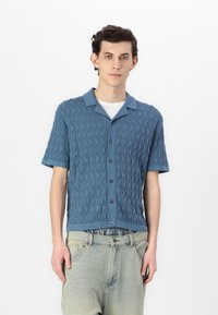 Short-sleeved, blue knit shirt with a wave pattern, collar, and button front. Worn with light-colored, loose-fitting jeans.