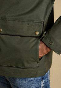 Nahaufnahme einer grünen Jackentasche mit Messing-Druckknöpfen, schwarzem Rand, "JOULES" geprägt und einer Hand, die teilweise in der Tasche steckt.