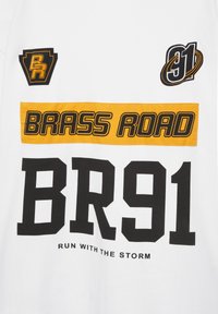 Tissu blanc avec des patchs noirs et jaunes indiquant "BRASS ROAD", "BR91" et "RUN WITH THE STORM", ainsi que des logos graphiques BR et 91.