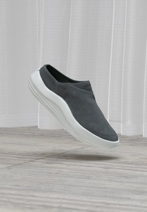 Chaussure à enfiler gris foncé avec une semelle blanche, flottant au-dessus d'un sol carrelé gris et de rideaux blancs en arrière-plan.