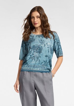 Kobieta z falującymi brązowymi włosami, ubrana w niebieską bluzkę z krótkim rękawem w kwiatowy wzór oraz luźne szare spodnie, stojąca z jedną ręką w kieszeni.