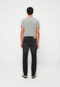 Un homme de dos portant un t-shirt gris, un jean noir avec des poches arrière, et des baskets blanches, se tenant devant un fond blanc uni.