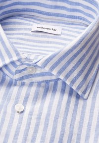 Gros plan d'une chemise habillée à rayures verticales bleues et blanches avec des boutons blancs et une étiquette "seidensticker" à l'intérieur du col.