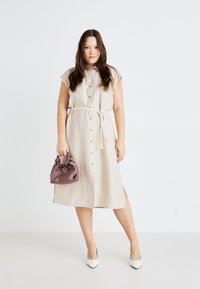 Vestido camisa de linho bege com mangas curtas, frente com botões, cintura com laço e aberturas laterais, combinado com uma bolsa de tecido roxa e saltos brancos.