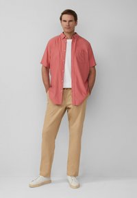 Koraalkleurig overhemd met korte mouwen en een borstzak, gecombineerd met beige broeken en witte sneakers, staand tegen een neutrale achtergrond.