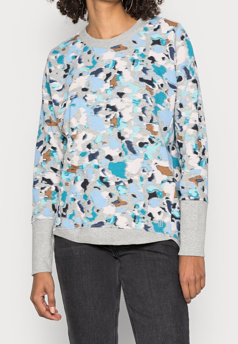 Femme portant un sweat-shirt à manches longues avec un motif abstrait bleu, gris et marron, avec des poignets et un ourlet gris clair.