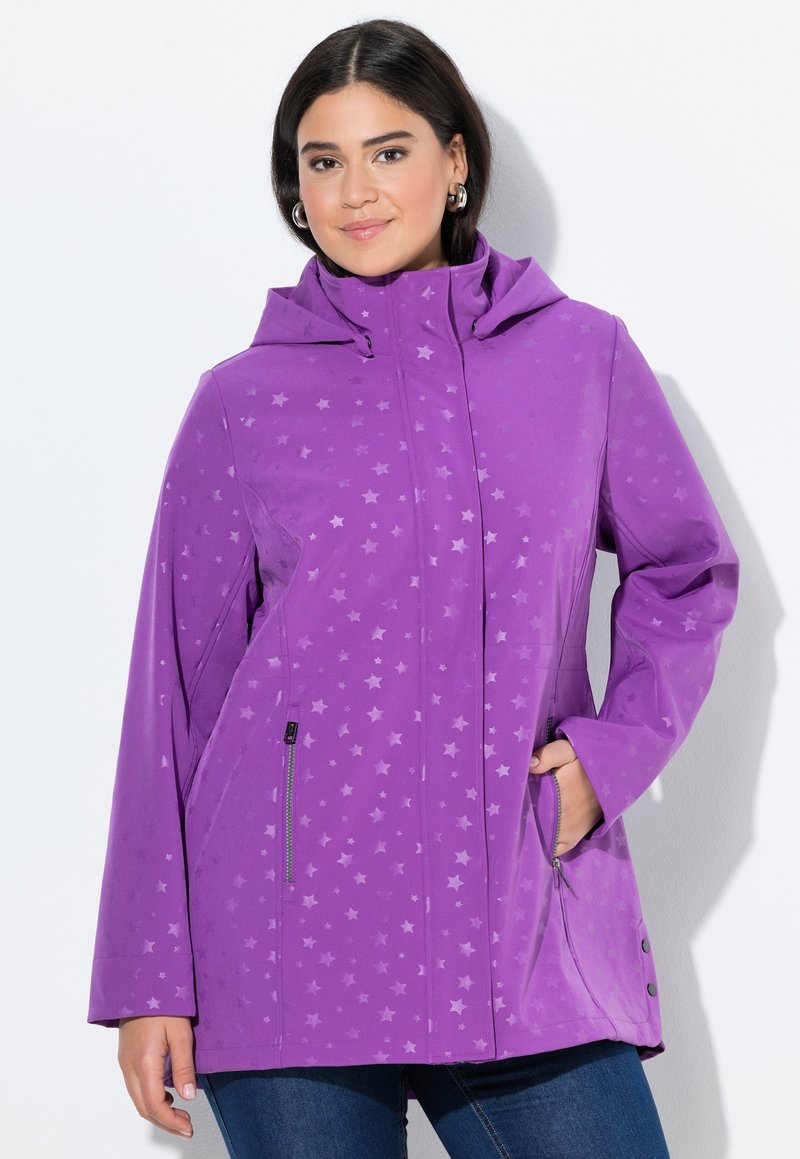 Donna che indossa una giacca a cappuccio viola con sottili motivi a stella, tasche con zip e jeans blu, in piedi su uno sfondo bianco.