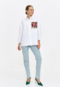 Biała koszula z guzikami i kolorową kieszonką w kwiaty, w połączeniu z jasnoniebieskimi dżinsami i zielonymi szpilkami. Gładka tekstura i luźny krój.