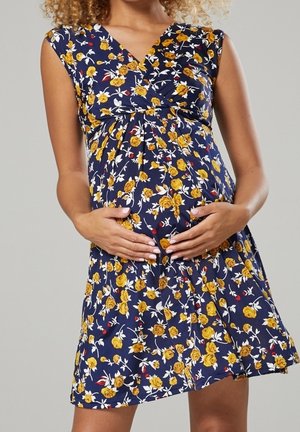 Gravid kvinde iført en mørkeblå kjole med gult blomsterprint, der blidt holder sin mave med begge hænder.