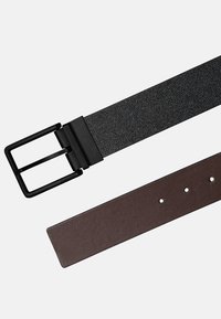 Cinturón reversible con tela negra texturizada por un lado y cuero marrón liso por el otro, con una hebilla de metal negro mate.