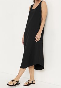 Fyrkantig svart klänning utan ärmar med en avslappnad passform, rundad halsringning och hög-låg fåll. Slät tyg kombinerat med platta svarta sandaler.