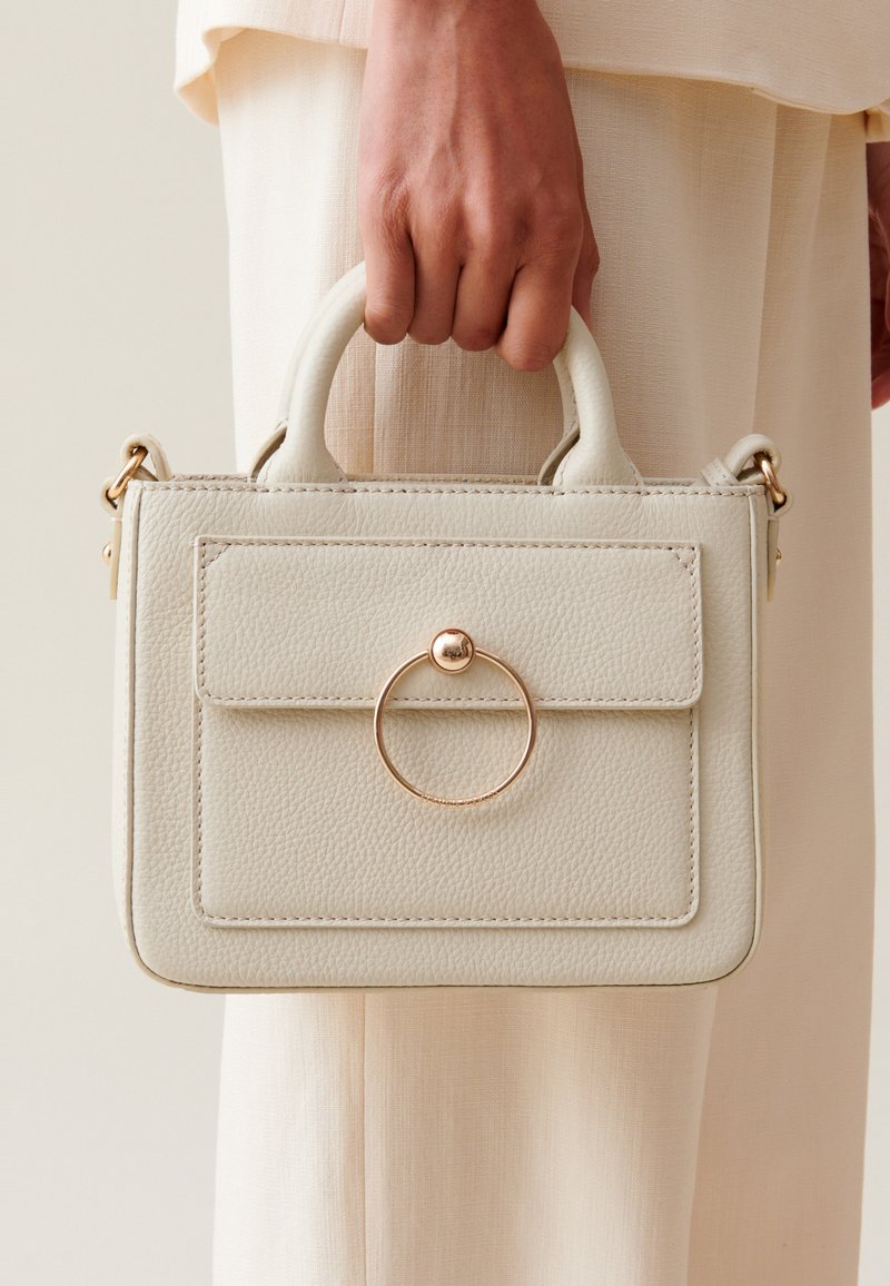 Sac à main en cuir crème avec un design structuré, une poche avant et un accent en métal doré en forme de cercle sur le rabat. Deux poignées supérieures.