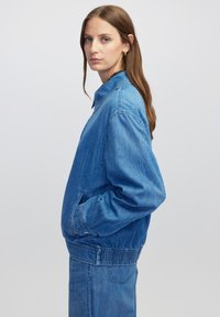 Vrouw met lang bruin haar, gekleed in een blauwe denimjack en bijpassende broek, in profiel staand met handen in de zakken van het jack tegen een witte achtergrond.