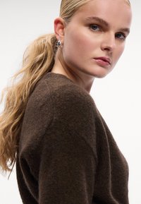Suéter marrón texturizado con cuello redondo y ligero detalle en los hombros, combinado con pendientes de estudio circulares, sobre un fondo blanco.