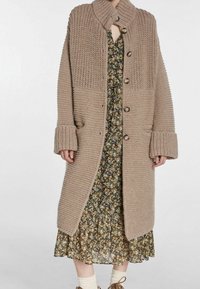 Beige neulottu neuletakki, jossa korkea kaulus, isot napit ja resorikuviointi. Varustettu sivutaskuilla ja leveillä hihansuilla, yhdistetty kukkamekkoon.