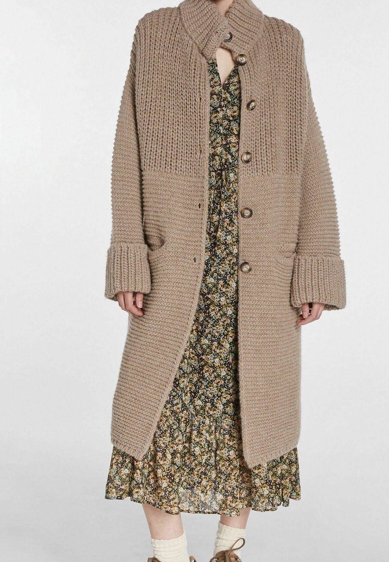 Beige neulottu neuletakki, jossa korkea kaulus, isot napit ja resorikuviointi. Varustettu sivutaskuilla ja leveillä hihansuilla, yhdistetty kukkamekkoon.