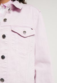 Jasnoróżowa dżinsowa kurtka z metalowymi guzikami i kieszenią z klapą na piersi, noszona na jasnoróżowej koszuli z kołnierzykiem.