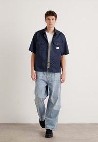 Kortermet marineblå skjorte med to brystlommer, grå innerventil, parret med lyseblå vide jeans og svarte sko. Enkel, avslappet stil.