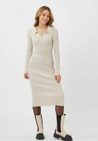 Abito beige a maniche lunghe con coste, colletto e scollatura a V. Indossato con stivaletti crema e neri, su uno sfondo bianco semplice.