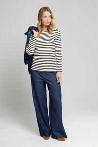 Stribet marineblå og hvid langærmet top parret med brede marineblå bukser, holdt med den ene hånd, mens den anden arm draperer en blå jakke.