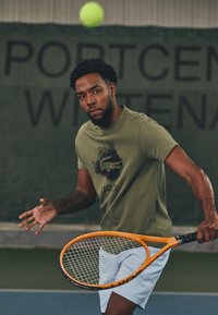 Green cotton t-shirt with a black graphic and text, paired with light blue shorts. Holding an orange tennis racket, focused on a green tennis ball.