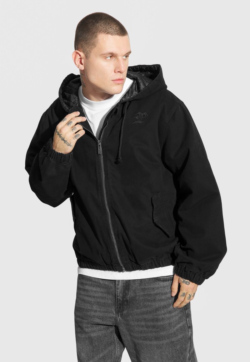 Czarna kurtka zapinana na zamek z kapturem, z elastycznymi mankietami i dołem, z delikatnym logo. Nosi się ją na białej koszuli i zestawia z szarymi dżinsami.