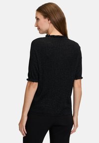 Sort bluse med korte ærmer og et tekstureret strikdesign, der har en flæsekrave og flæser ved ærmerne, parret med tætsiddende sorte bukser.