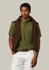 Zelená polo tričko s golierom, s logom, hnedý pletený sveter prehodený cez ramená. Model má na sebe svetlé nohavice.