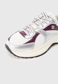Chaussure de sport argentée et blanche avec matière en mesh, accents texturés, et logo violet. Semelle en caoutchouc à design stratifié avec lacets.