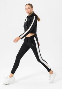 Felpa nera corta con strisce bianche, abbinata a joggers neri con pannelli laterali bianchi e sneakers bianche.