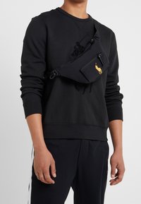 Svart nylonaxelväska med en framfickan med dragkedja, prydd med en gyllene logotyp. Bärs diagonalt över en svart sweatshirt och byxor.