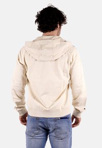 Felpa con cappuccio beige dal tessuto liscio, con polsini e orlo elasticizzati, caratterizzata da un design triangolare sul retro. Indossata con jeans blu.