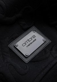 Tissu noir texturé avec une étiquette rectangulaire noire cousue, embossée du texte « ombre menswear » et quatre rivets aux coins.