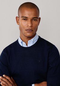 Marineblauwe crewneck sweater gelaagd over een lichtblauw overhemd met kraag. De sweater heeft lange mouwen en een subtiele geborduurde logo op de borst.